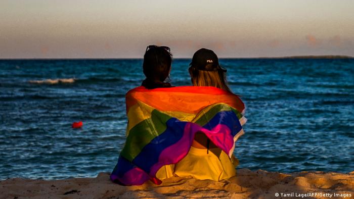Two women sit at the beach wrapped in a rainbow flag 