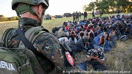Ein Soldat bewacht zahlreiche Flüchtlinge, die auf der Erde im Gras hocken und die Hände über dem Kopf halten