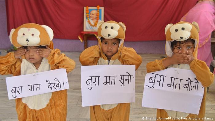 Drei kleine Kinder stehen in Affenkostümen in einer Reihe nebeneinander und haben weiße Blätter mit Sanskrit-Schrift vor ihrer Brust hängen.