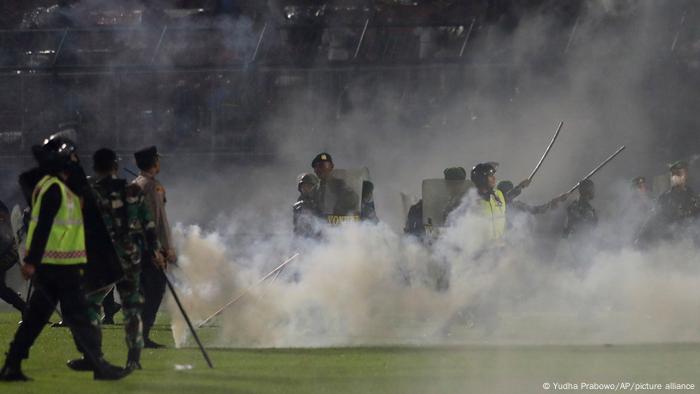 Police officers armed with batons stand between clouds of tear gas on the lawn of Kanjuruhan Stadium