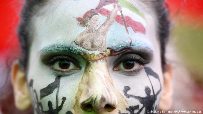 Close up of a woman's face painted with the iconic 'Marianne' of France waving an Iranian flag and prominent figures protesting.
