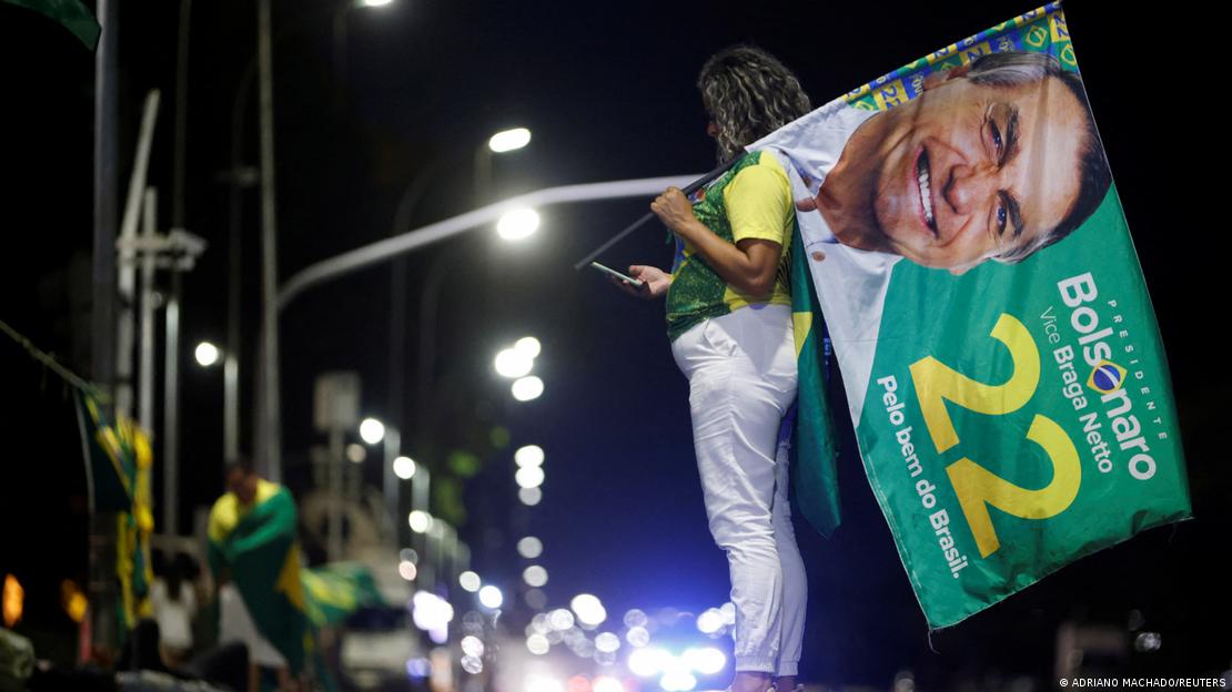 Mulher com camiseta verde e amarela e calça branca olha celular enquanto segura bandeira com os dizeres 