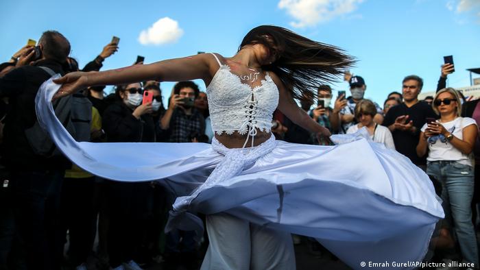 Crowds of people film a woman with long black hair, dressed in a white crop top and white skirt and twirling around like a dervish.
