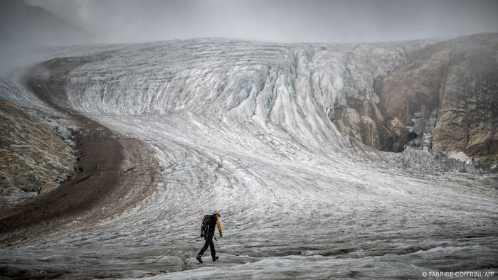 Eine Person mit Rucksack und Messgerät schreitet über einen schneefreien Gletscher, an den Seiten ist der Fels zu sehen