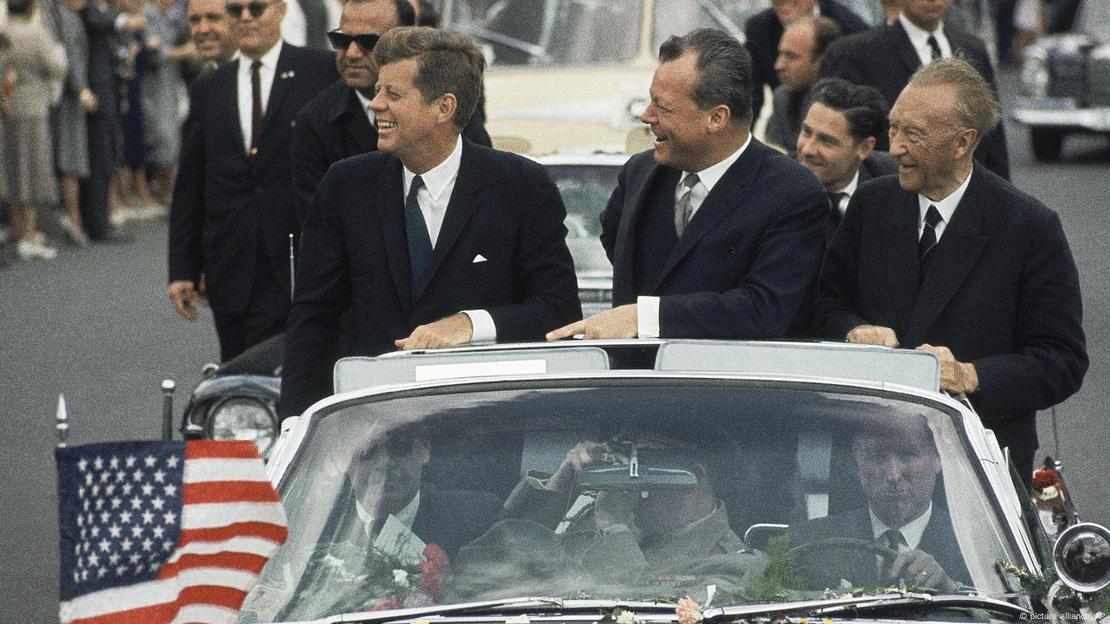 John F. Kennedy, Willy Brandt i Konrad Adenauer stoje u automobilu s otvorenim krovom i zastavicom SAD-a i smiješe se ljudima John F. Kennedy, Willy Brandt i Konrad Adenauer stoje u automobilu s otvorenim krovom i zastavicom SAD-a i smiješe se ljudima
