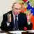 O presidente da Rússia, Vladimir Putin, está sentado e de braços abertos, como se estivesse questionando. Ele usa terno e gravata escuros, com camisa branca. Ao fundo, há uma bandeira da Rússia.
