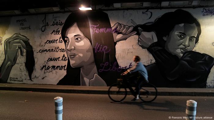 Une femme fait du vélo devant une peinture murale signée par Clacks-one et Heartcraft_Street art, représentant des femmes se coupant les cheveux pour montrer leur soutien aux manifestants iraniens 