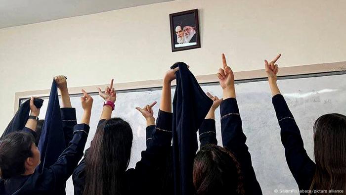 Female students, shot from behind in the classroom, show obscene gestures in front of portraits of the religious leaders Khomeini and Khamenei