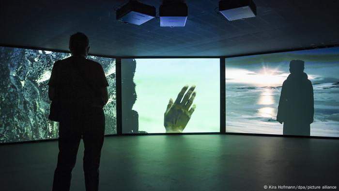 Eine Person steht vor einem dreigeteilten großen Bildschirm, der Folgendes zeigt: Eine Person, die am Meer steht, eine Hand und Strukturen. Eine Person steht vor einem dreigeteilten großen Bildschirm, der Folgendes zeigt: Eine Person, die am Meer steht, eine Hand und Strukturen.
