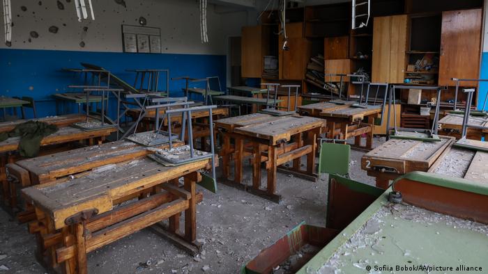 Ukraine Krieg | Schule in der Region Charkiw. Das Foto zeigt einen Klassenraum nach einem russischen Angriff in der Nähe des Schulgebäudes. Auf den Holztischen, auf denen die Stühle hochgestellt sind, liegt Staub und Schutt. Die Wand des Klassenzimmers ist von Einschusslöchern durchsiebt. Von der Decke hängen Kabel.