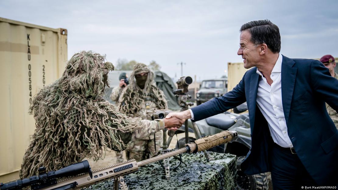 Mark Rutte, in an open-collared white shirt and dark blue suit, shakes hands with a soldier in a shaggy camouflage outfit
