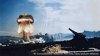 Un cañón se ve mientras al fondo se eleva la explosión atomíca con su característico humo en forma de hongo.