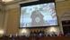 Sala da comissão com congressistas sentados em mesa e telão com imagem de Trump discursando Sala da comissão com congressistas sentados em mesa e telão com imagem de Trump discursando
