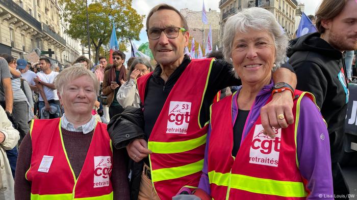 Three older people standing amid a crowd of protesters wearing red vests with yellow fluorescent stripes 