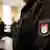 A guard in a courtroom stands in the foreground, dressed in black with a patch on his upper arm that reads "Justice" in German and the logo of the Hanseatic Higher Regional Court. His face is not seen. In the background, three people stand, out of focus 