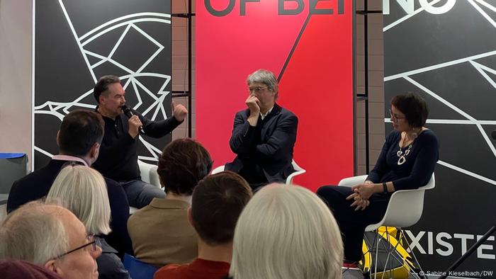 Juri Andruchowytsch auf einem Podium der Frankfurter Buchmesse mit zwei weiteren Personen. Er spricht ins Mikrofon. 