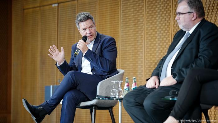 Bundeswirtschaftsminister Robert Habeck und BDI-Präsident Sigfried Russwurm sitzen auf dem BDI-Rohstoffkongress auf zwei Stühlen im Gespräch nebeneinander. Habeck hält ein Mikrofon in der Hand und spricht. Die beiden Männer tragen dunkle Anzüge. Auf einem kleinen Tisch zwischen den Stühlen stehen Gläser und Flaschen. Bundeswirtschaftsminister Robert Habeck und BDI-Präsident Sigfried Russwurm sitzen auf dem BDI-Rohstoffkongress auf zwei Stühlen im Gespräch nebeneinander. Habeck hält ein Mikrofon in der Hand und spricht. Die beiden Männer tragen dunkle Anzüge. Auf einem kleinen Tisch zwischen den Stühlen stehen Gläser und Flaschen.