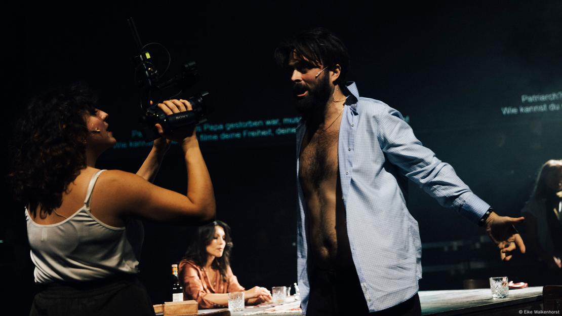 A woman in a tank top holds a camera at a man with his bare chest showing.