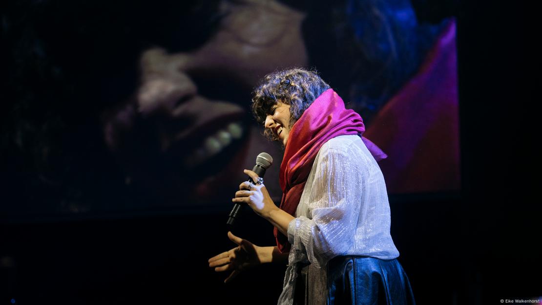 A woman wearing a red piece of fabric and singing into a microphone with an image of her face projected on a screen behind her.