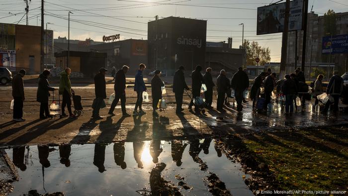 Ukraine | Krieg | Zerstörte Trinkwasserversorgung in Mykolajiw. Auf dem Foto sind Menschen zu sehen, die mit leeren Kanistern in einer Schlange stehen. Im Hintergrund sind Gebäude zu sehen, im Vordergrund eine große Wasserpfütze. Die Aufnahme ist gegen die Sonne gemacht, deswegen sind die Menschen vor allem als Schatten zu sehen. Ukraine | Krieg | Zerstörte Trinkwasserversorgung in Mykolajiw. Auf dem Foto sind Menschen zu sehen, die mit leeren Kanistern in einer Schlange stehen. Im Hintergrund sind Gebäude zu sehen, im Vordergrund eine große Wasserpfütze. Die Aufnahme ist gegen die Sonne gemacht, deswegen sind die Menschen vor allem als Schatten zu sehen.