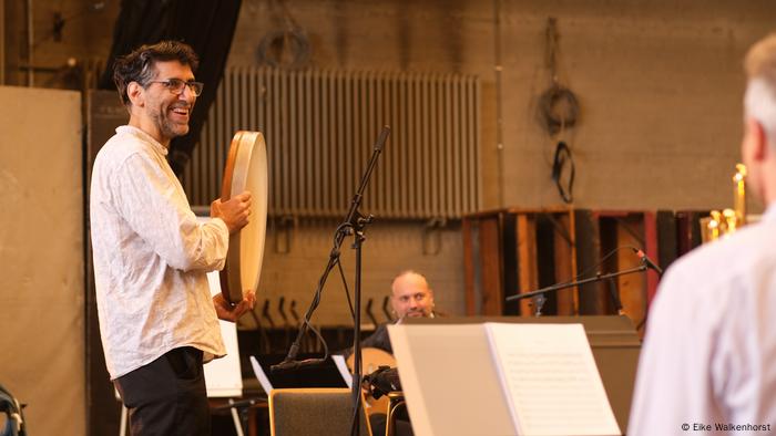 Lächelnd schlägt Keyvan Chemirani eine Trommel, während zwei weitere Musiker ebenfalls proben. Lächelnd schlägt Keyvan Chemirani eine Trommel, während zwei weitere Musiker ebenfalls proben.