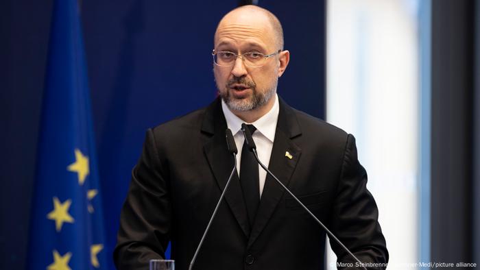 Hombre con barba y gafas de traje hablando ante dos micrófonos. 