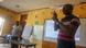 Dr. Kodwo Boateng (links, im Hintergrund) und Frederick Anderson stehen in einem Klassenraum. Anderson erklärt etwas in Gebärdensprache. Im Hintergrund wird eine Präsentation mit einem Beamer auf ein Whiteboard geworfen.