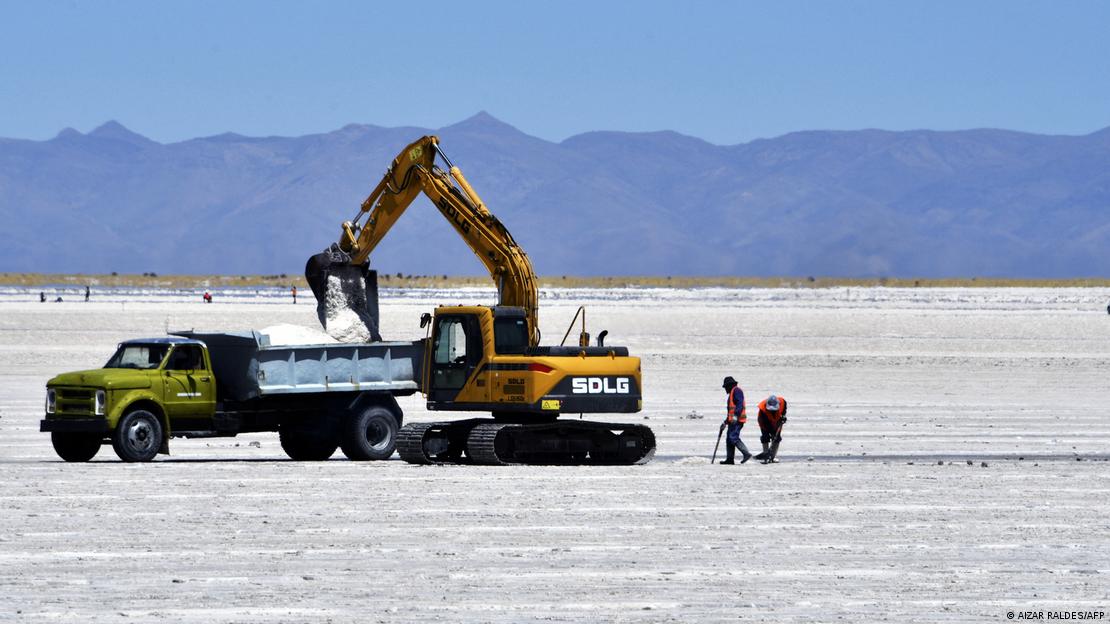 Extra&ccedil;&atilde;o de l&iacute;tio na Argentina; retroescavadeira despeja terra branca em uma ca&ccedil;amba de caminh&atilde;o. Dois homens seguem atr&aacute;s do maquin&aacute;rio. O cen&aacute;rio &eacute; uma salina, de solo muito branco, com montanhas e c&eacute;u azul ao fundo.