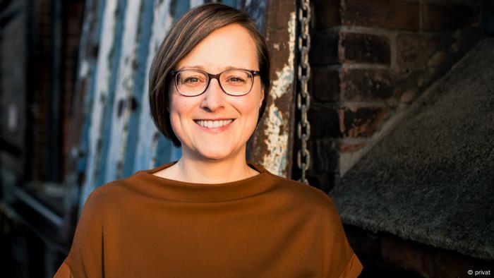 Journalism Professor Christina Elmer smiles into the camera. In the head shot, she wears a light brown sweatshirt and dark-framed glasses.