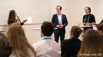 Two panelists and a moderator seen from behind the heads of listening conference participants. Christina Elmer holds a microphone in her hand and is about to answer on the right side of the image.