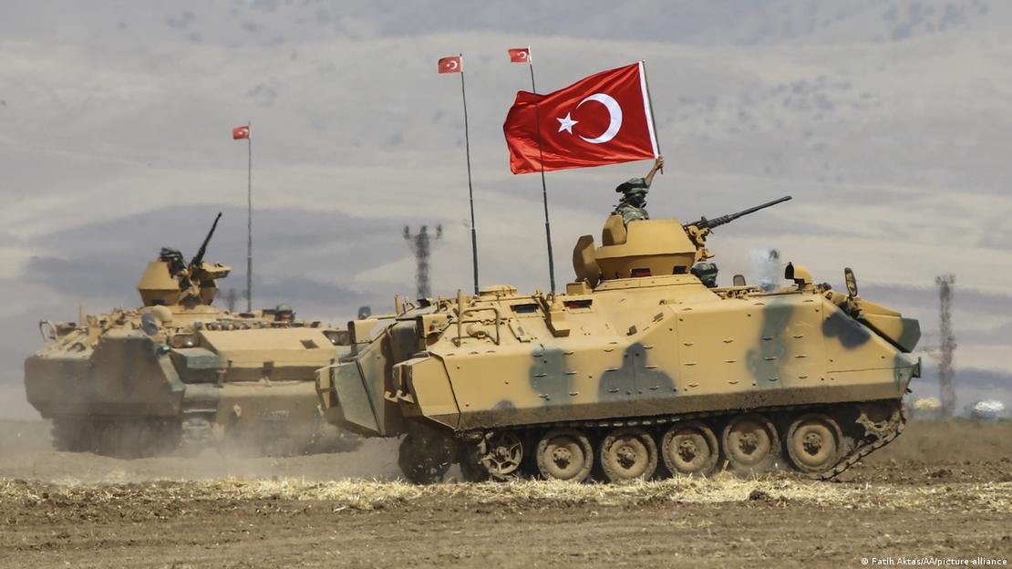 A tank hurtles across a stoney ground while a soldier holds a Turkish flag