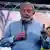 O presidente eleito Luiz Inácio Lula da Silva veste camisa jeans e olha compenetrato para uma plateia, para a qual fala com um microfone. Ele segura o microfone com a mão direita e ergue o dedo indicador com a mão esquerda.