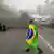 A protester at a roadblock walks with a Brazilian flag draped over their back. In the background, something is on fire
