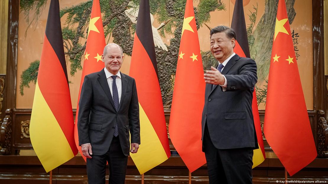 Njemački kancelar Olaf Scholz i kineski predsjednik Xi Jinping ispružene ruke prema kameri a iza njih po tri njemačke i kineske zastave