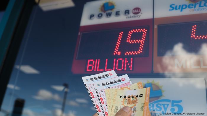 Una mano muestra billetes de lotería junto al anuncio del bote acumulado.