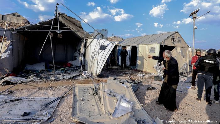 Ein Mann steht vor einem zerstörten Haus im Flüchtlingslager und zeigt mit dem Finger darauf