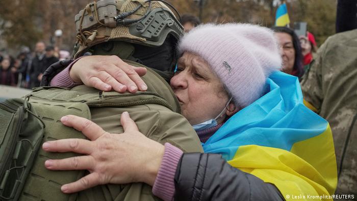 Alte Frau mit umgehängter ukrainischer Flagge umarmt Soldaten