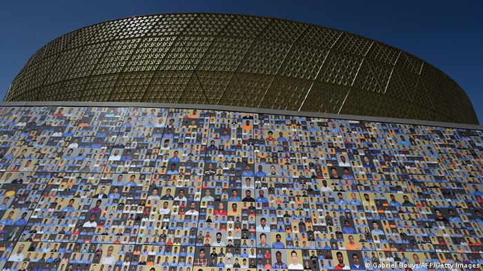 Eine Wand mit Hunderten bunten Fotos, dahinter erhebt sich das glänzend sandfarbene Stadion