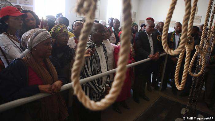 Eine Besuchergruppe steht vor der Absperrung zu einer ehemaligen Hinrichtungsstätte in Südafrika, hinter der mehrere Galgenseile hängen 