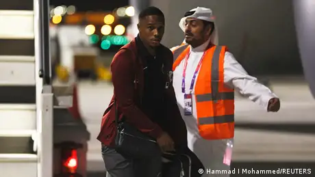a Qatari football player, wearing a brown sweater walking past a security staff.