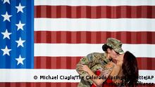 Delante de la imagen de la bandera estadounidense, una soldado besa a su esposa. 