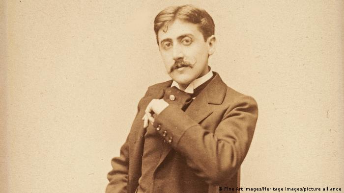 Marcel Proust im Anzug. Er schaut mit erhobenem Arm in die Kamera.