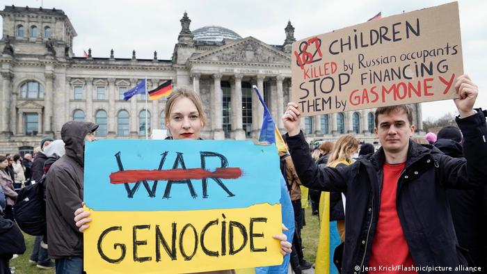 Një grua e re mban në duar një poster me ngjyrat ukrainase blu dhe të verdhë përpara ndërtesës së Reichstag në Berlin. Fjala Luftë është e kryqëzuar me të kuqe me Genocid poshtë. Pranë tij është një djalë i ri që mban lart një pankartë. Ndër të tjera thuhet: 167 fëmijë të vrarë nga pushtuesit rusë