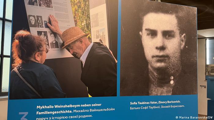 Zwei Fotos auf einem Aufsteller: Ein älterer Mann steht an einer Ausstellungstafel und berührt sie mit der Hand. Neben ihm steht eine Frau. Rechts ein Porträtbild