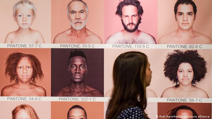 Ein Frau läuft an einem Bild mit Porträts von Menschen verschiedener Hautfarben vorbei, welches die Hauttypen von Menschen in Pantone-Farben aufschlüsselt.