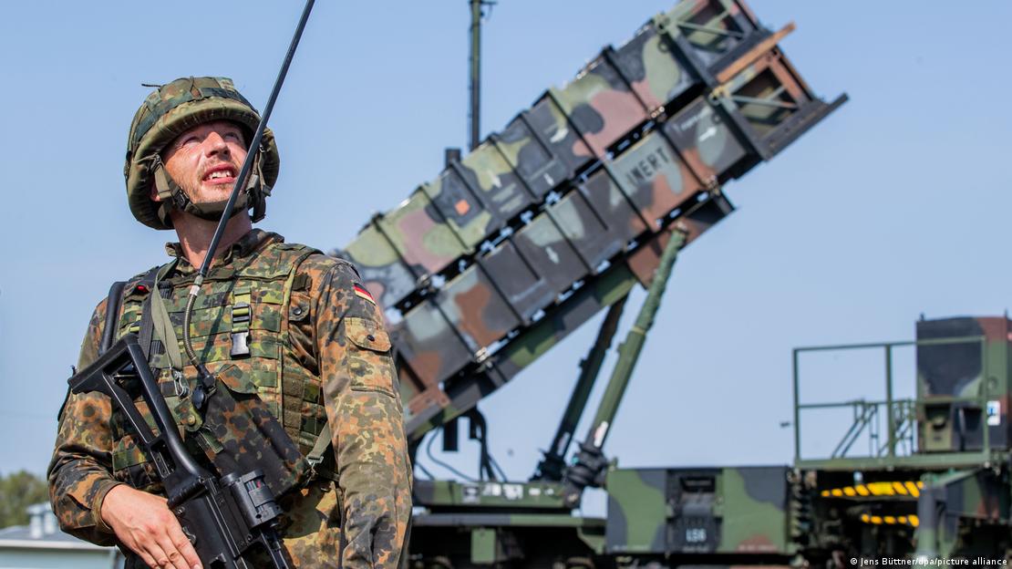 A German soldier in uniform stands in front of a mobile Patriot missile launching station, looking up at the sky