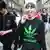 A young woman in a black and green T-shirt referencing marijuana addiction and the German sportswear company Adidas, licks a bright green lollipop as she smiles into the camera while walking along a crowded street