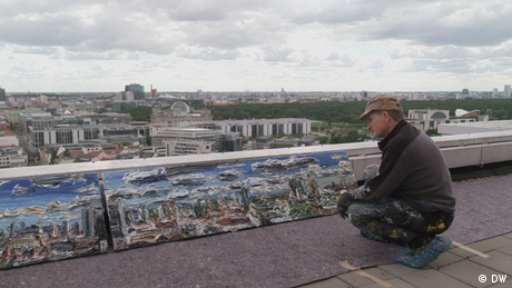 Der Maler Christopher Lehmpfuhl hockt auf dem Dach eines Hauses und betrachtet seine Gemälde (Quelle: DW)