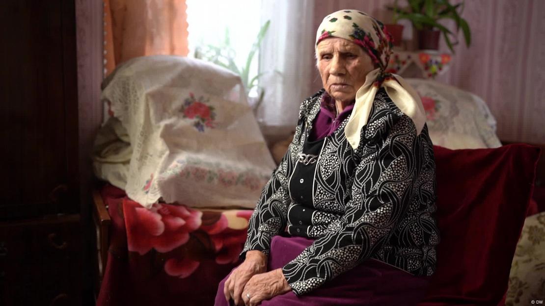 Liubov Yarosh, a 102-year-old Ukrainian woman wearing a headscarf and a black and white patterned blouse, sits in her home