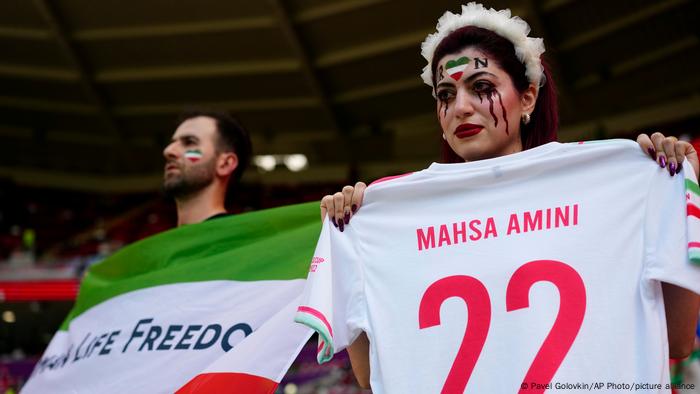 Dos fans en las gradas. A la dercha, una mujer sostiene una camiseta de fútbol con el número 22 y el nombre de Mahsa Amini.